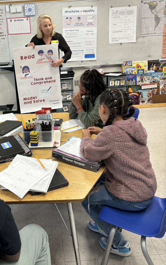 A teacher is holding a Structures of Equality poster and referencing it during small group work with students.